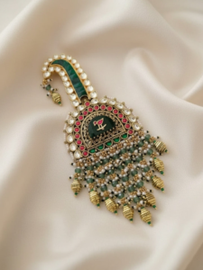 Ornate gold pendant featuring green and red stones, a pearl-bordered rim, and dangling bead fringe on ivory satin.