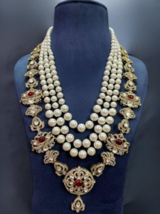 Layered pearl and gold necklace set on a dark display bust, featuring multiple strands and red gemstones at the center and sides.