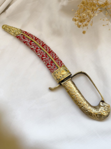 Ornate dagger with a gold-hued carved handle and a red embroidered sheath resting on white fabric with dried flowers nearby.