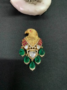 Gold eagle-shaped brooch with pink accent stones, clear crystals, and dangling green emerald teardrops on a dark background.